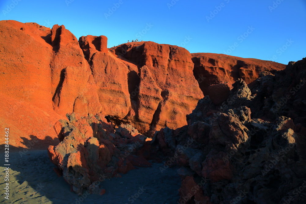 Obraz premium The rocky and volcanic coast in El Golfo, Lanzarote, Canary Islands, Spain, located next to Lago Verde, with at the top people enjoying the view from the mirador (viewpoint) del Charco de Los Clicos