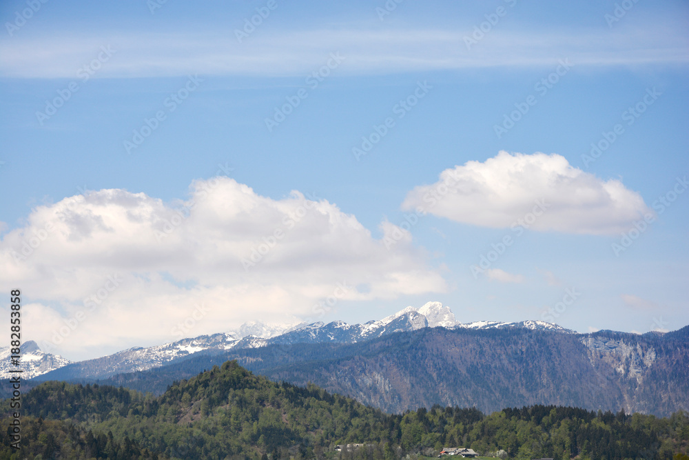 Fototapeta premium A wide landscape view of the distant snow capped peaks of the Julian Alps rising above the lush green forests and hills of the Gorenjska region in Slovenia
