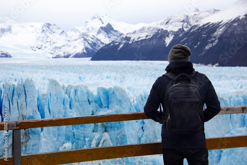 Tourists standing on a metal observation deck look out at the massive blue ice face of Perito Moreno Glacier beneath snow-capped peaks in Patagonia, Argentina.