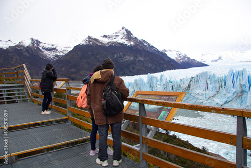 Tourists standing on a metal observation deck look out at the massive blue ice face of Perito Moreno Glacier beneath snow-capped peaks in Patagonia, Argentina.