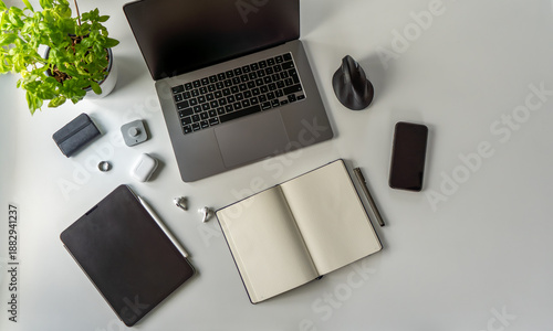 Contemporary flat lay of creative workspace including open dotted journal notebook, smart ring wearable, ergonomic mouse, laptop, tablet, phone and basil plant. Clean modern office setup.