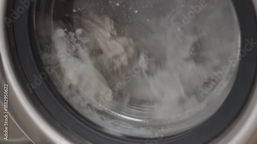 Close up transparent washer door showing soapy water and fabric tumbling inside, droplets streaking glass, stainless rim in frame,clean household textiles swirling under foam