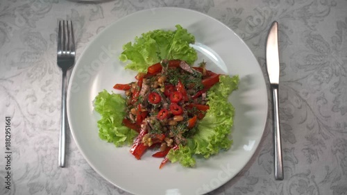 Top view of a fresh, spicy georgian salad with meat, beans, walnuts, and hot peppers on a lettuce leaf, slowly rotating on a white plate