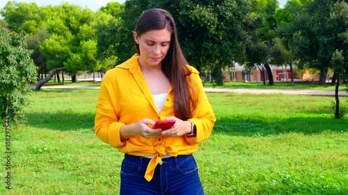 Concerned woman wearing yellow shirt looking at smartphone outdoors with hand on forehead in park, concept of stress, bad news, frustration, disappointment, communication, technology, emotion.