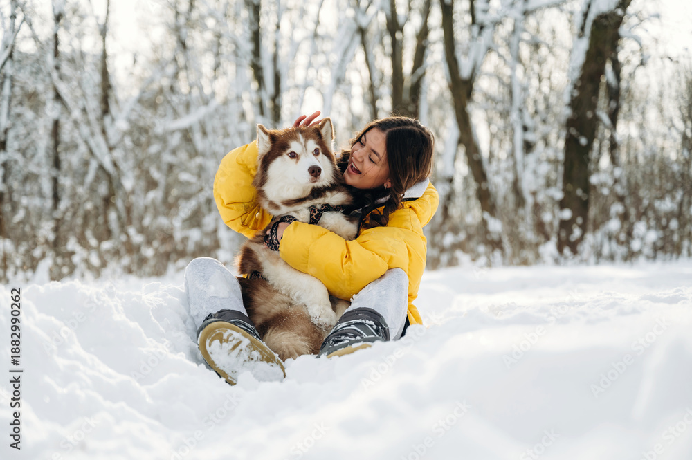 Obraz premium Young woman in a yellow puffer jacket is sitting in the snow while holding a brown and white husky dog, surrounded by snow-covered trees in a winter landscape