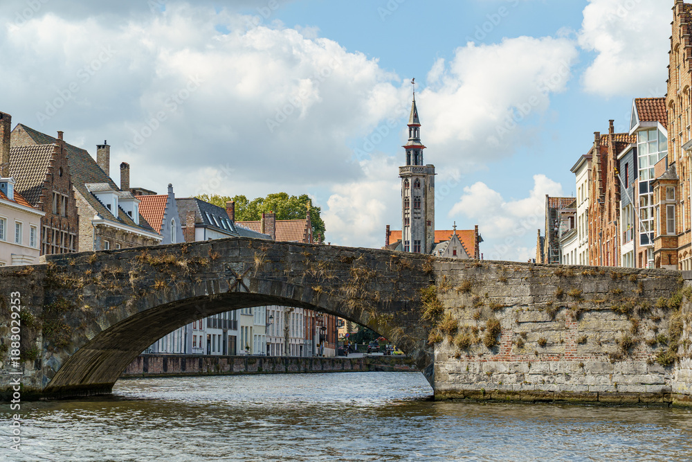 Fototapeta premium A stone bridge over a canal in Bruges, Belgium. A tall Gothic church tower is visible in the background