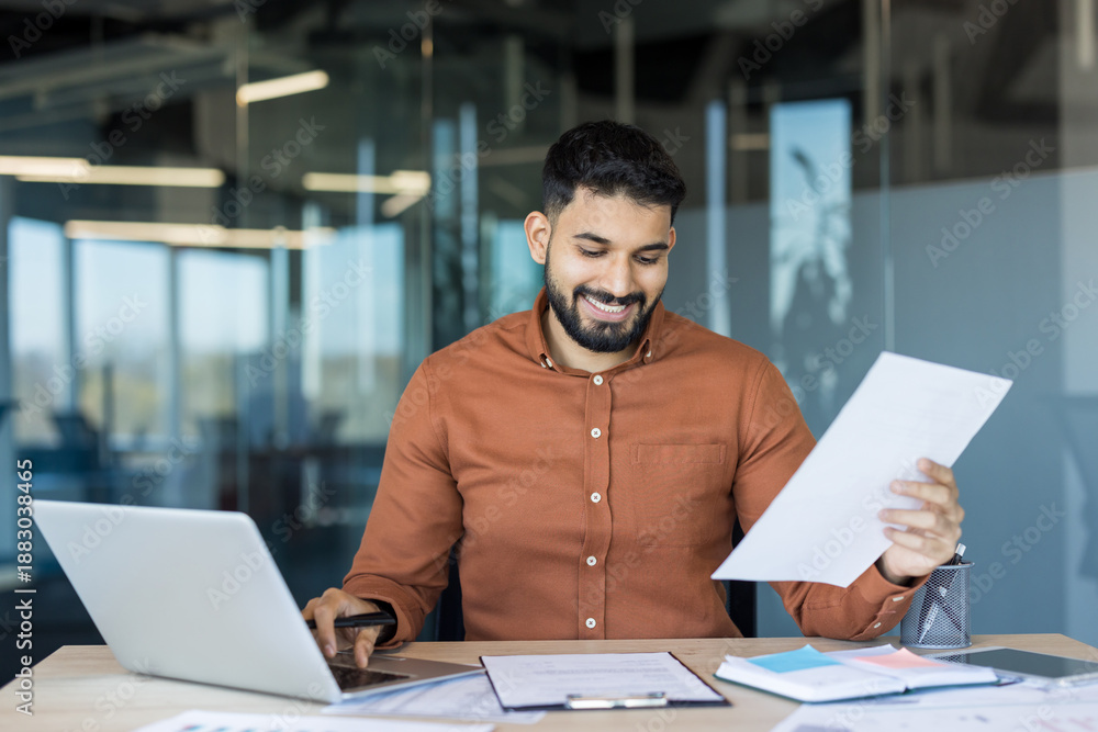 Obraz premium Young confident bearded man holding documents and typing on laptop, reviewing paperwork and analyzing data while sitting at a desk in a contemporary clear glass office environment