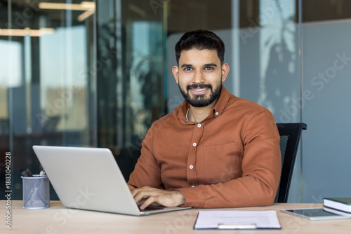 Young professional man with a beard sitting at his desk, typing on a laptop, and looking confidently at the camera, portraying business, success, and leadership in a modern office environment