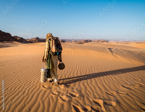 Nomadic traveler carrying traditional baskets walking across sand dunes in a vast desert landscape.