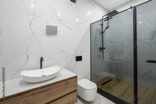 Modern minimalist bathroom with white marble walls, glass shower enclosure, wooden vanity, oval sink, wall-mounted toilet, sleek black fixtures, light wood flooring.