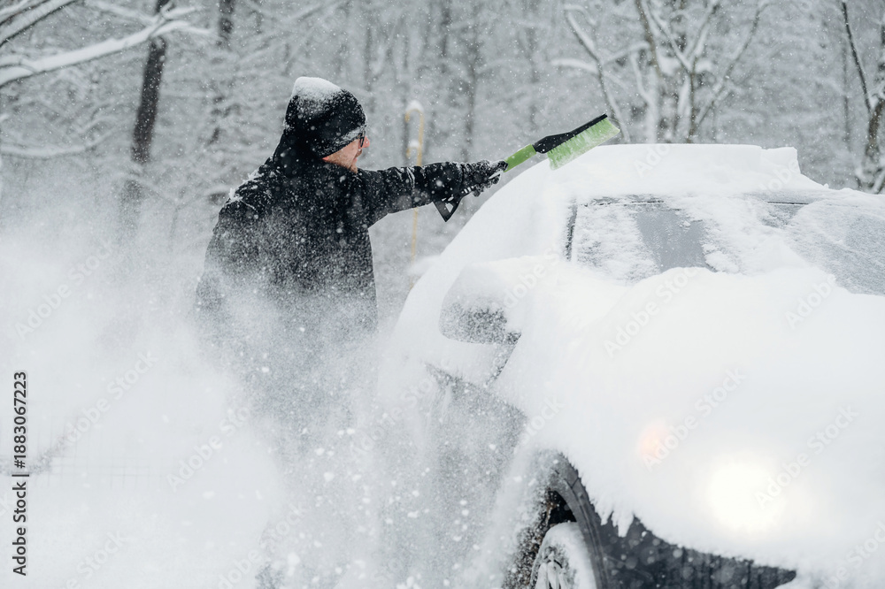 Fototapeta premium Person in black winter attire is clearing snow from a car parked in a snowy landscape, trees covered in snow visible in the background during winter weather conditions