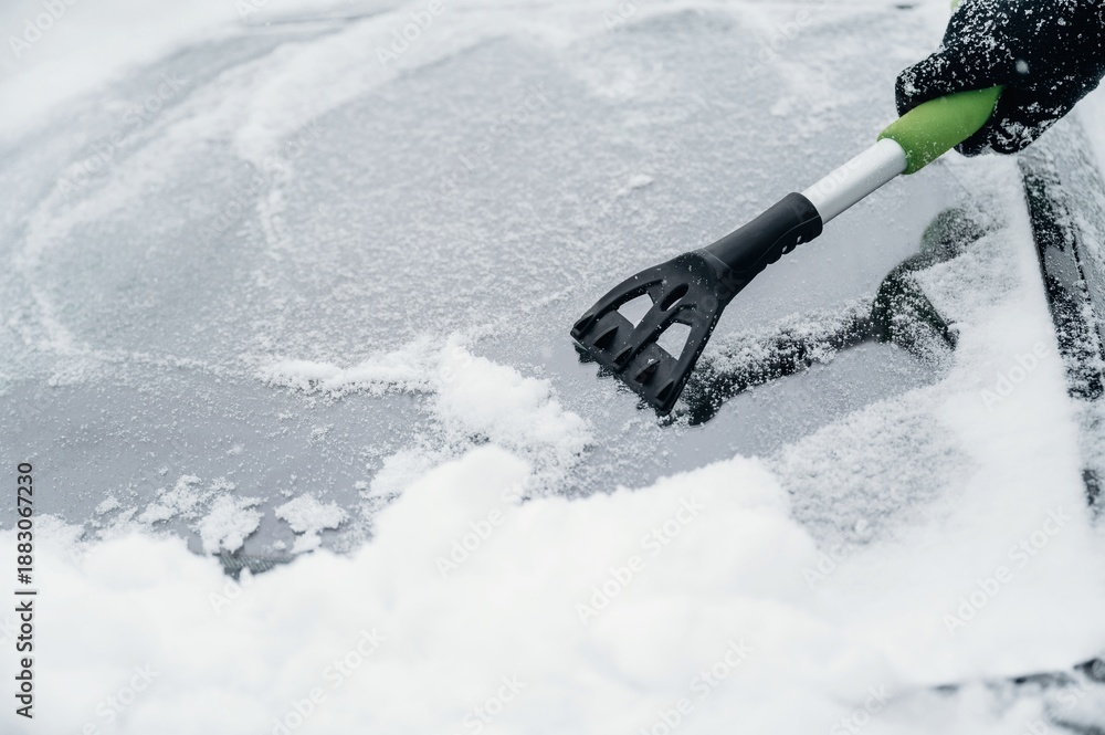 Fototapeta premium Person using a black and green snow scraper to remove snow from a car windshield covered in white snow during winter weather conditions in a cold environment
