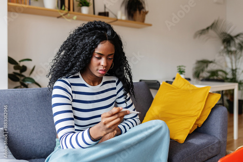 Young sick Latin American woman measuring fever with thermometer sitting on sofa at home