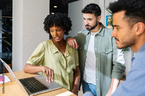 Diverse coworkers collaborating around laptop in creative office. Multicultural team analyzing information together. Innovation and teamwork concept.