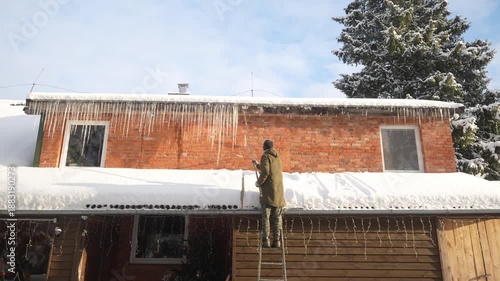 Man stands on the ladder and knocks down the icicles form the roof. Winter household work. Large icicles on the house.