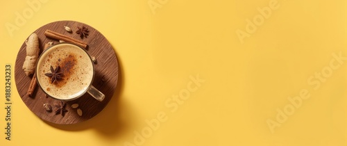 Traditional Indian masala chai latte in a glass cup featuring milk, spices, and herbs, placed on a wooden board on a yellow background with strong shadow, top view and blank space for text