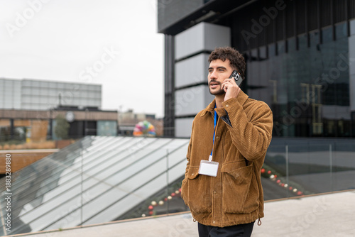 Young man talking on smartphone, wearing corduroy jacket and id badge in urban business area