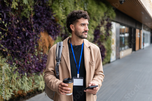 Young professional man commuting to work, walking with coffee and smartphone on urban street with id badge