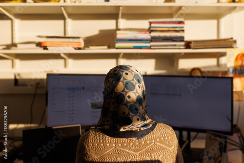 A Muslim woman seen from the back, working on two computer screens with book shelves in the background.