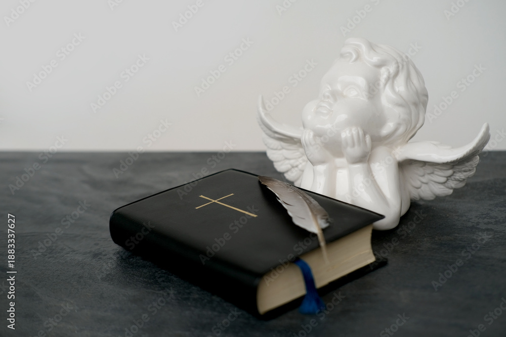 Obraz premium old black book, family bible in dark cover, white ceramic figurine of angel with wings, goose feather, concept of intermediary between God and world, eternal Christian values, unity of people in faith