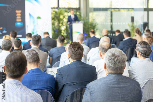 Business audience attending corporate keynote presentation, professional conference seminar with speaker at podium in modern event venue