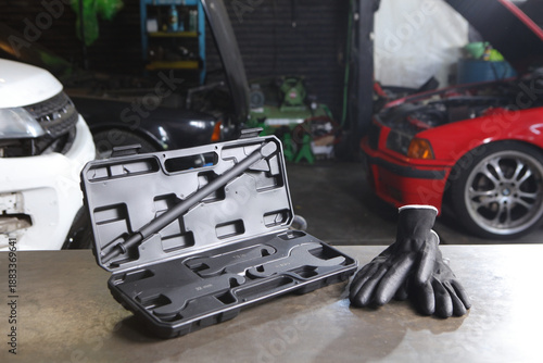 Quadro em tela An open black plastic tool case containing specialized automotive wrenches and a long metal rod sits on a workbench next to a pair of black work gloves in a garage with cars