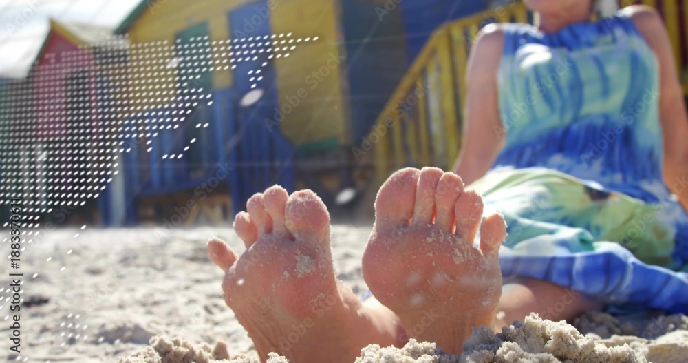 Fototapeta premium Relaxing bare feet sinking into granular sand at beach, showing blue-green sundress and huts