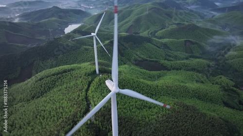 White wind turbines standing on green hills with dense forest and natural landscape captured from aerial view