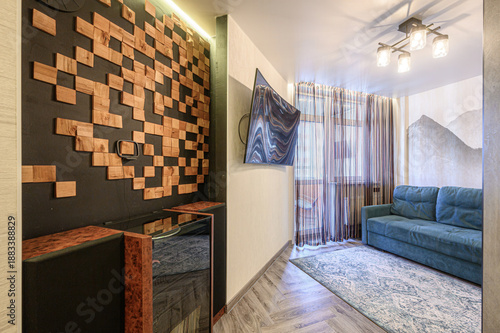 Modern living room with wooden mosaic wall, dark wooden cabinet, teal sofa, patterned rug, sheer curtains, wall-mounted TV, ceiling light fixture, mountain-themed wall art.
