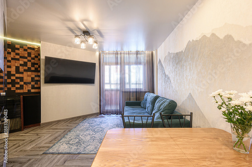 Modern living room with teal sofa, wooden dining table, wall-mounted TV, sheer curtains, mountain mural, patterned rug, ceiling light fixture, vase with white flowers.