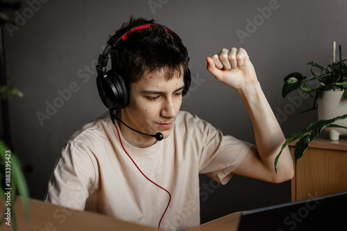 A guy in a beige T-shirt with his hand up and pimples on his face is sitting at a table with headphones connected to a laptop. streams, games, and online learning. preparation for exams. vertical phot
