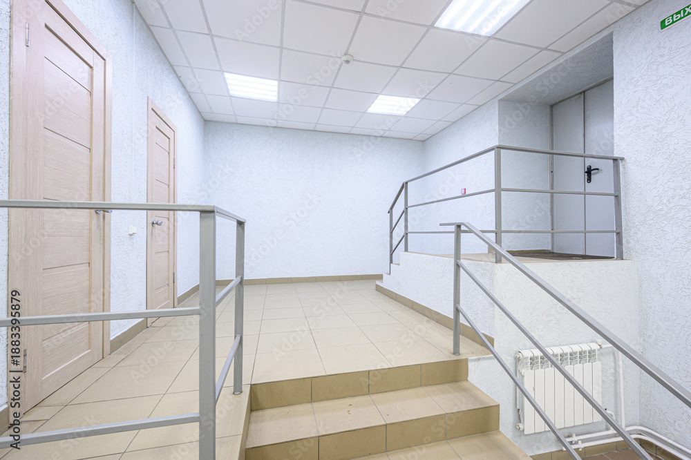 Obraz premium clean, minimalist hallway with beige tiled floor, white textured walls, metal railings, wooden doors, staircase, recessed ceiling lights, and white radiator. Functional, uncluttered space.