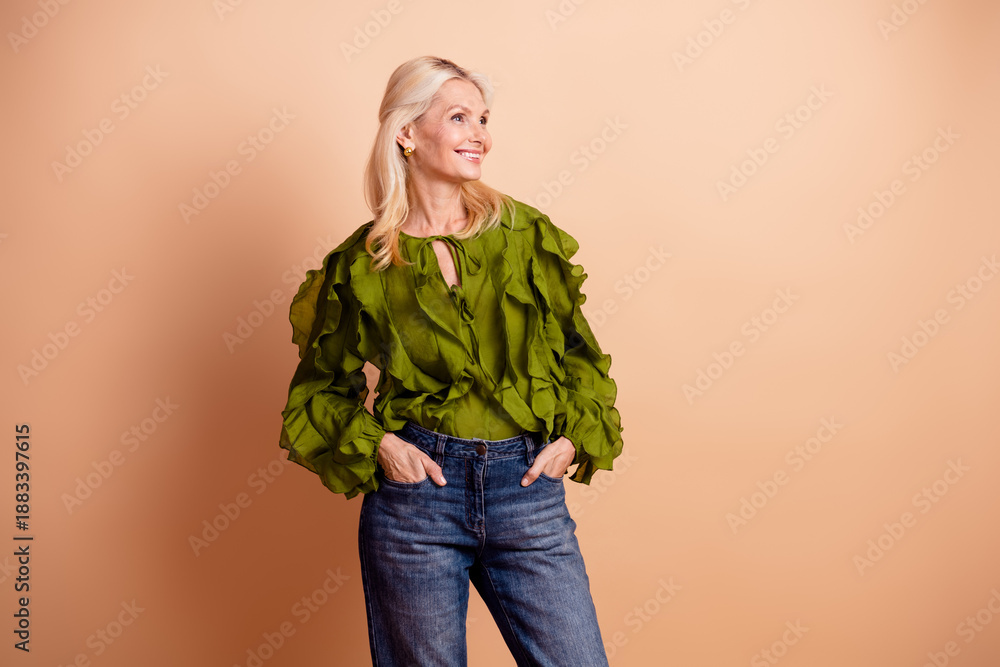 Fototapeta premium A mature beautiful female woman with blonde hair wearing a green blouse and jeans stands confidently against a beige background