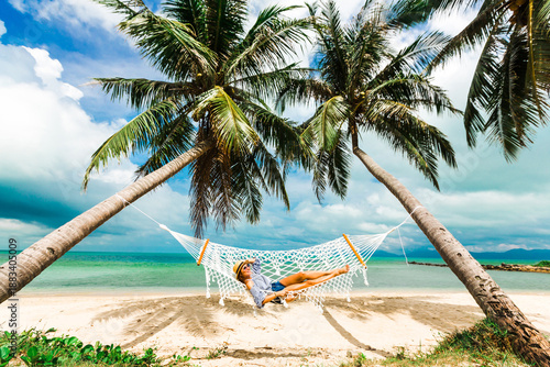 Happy beautiful girl in a straw hat and shorts, striped t-shirt, lying on a beach hammock between two palm trees, on the seashore of a tropical island, traveling summer vacation