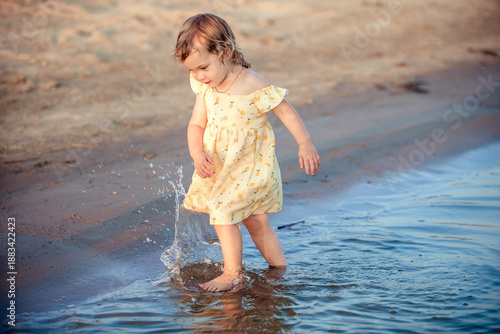 Fotografija 3 year old blonde girl running along the river bank, splashing water on a summer
