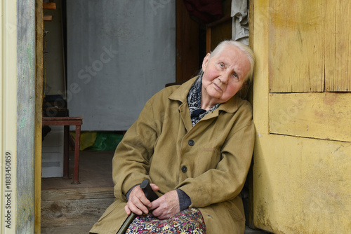 A sad elderly farmer woman with a walking stick is sitting on the porch of her old village house. Loneliness and poverty.