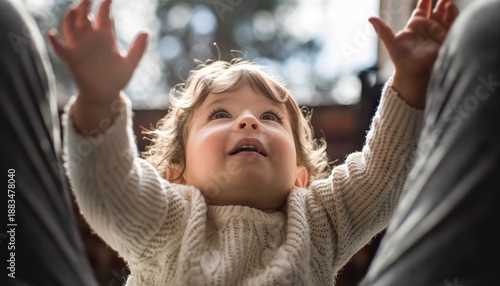 Happy toddler with curly brown hair reaches up with both hands while looking up with joy, wearing a cozy knitted sweater in a backlit scene with a soft focus background