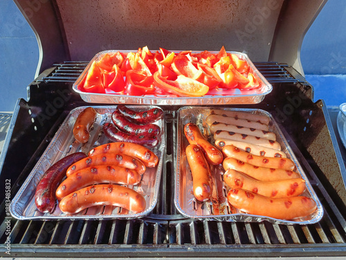 Grilling is the process of roasting vegetables and meats on the grill. Red bell peppers and various types of sausages and hot dogs with grill marks are cooked on aluminum baking sheets.