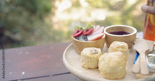 Delicious scones and fresh fruit platter served on a wooden tray with jam and drinks in a serene outdoor garden setting, evoking a relaxing and wholesome brunch experience.