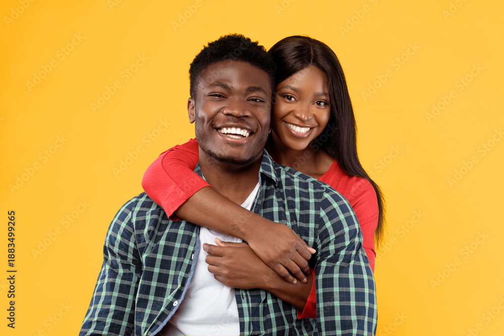 Fototapeta premium A man and a woman embrace each other while smiling. They stand in front of a bright yellow background. The man wears a checkered shirt, and the woman wears a red outfit.
