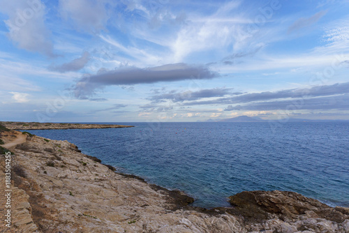 A high-angle, sun-drenched panoramic view of the pristine turquoise waters of Spiaggia di Cala Minnola on Levanzo Island, Sicily, with coastal buildings and the distant island of Favignana visible on 