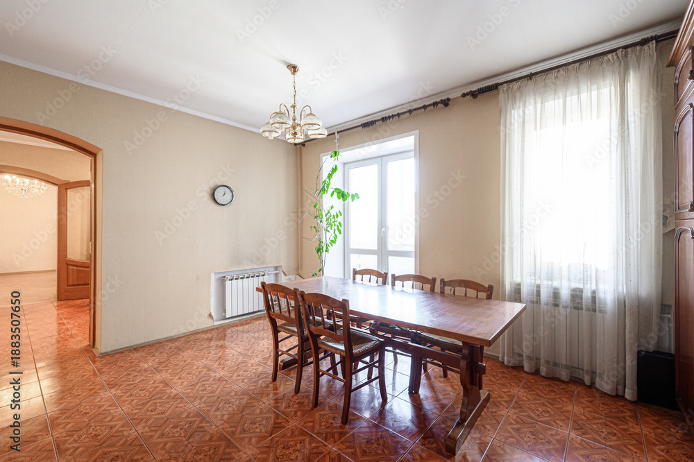 Fototapeta premium Spacious dining room with wooden table and chairs, chandelier, sheer curtains, large window, terracotta tiled floor, wall clock, radiator, arched doorway, potted plant. Light, airy ambiance.