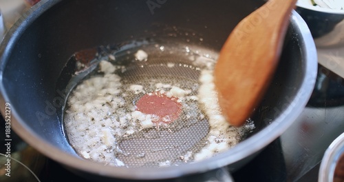 Chopped garlic and a red spice being sautÃ©ed in a hot frying pan with bubbling oil, stirred by a wooden spatula, capturing the vibrant essence of home cooking preparation.