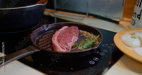 Tableau sur toile Fresh steak searing in a cast iron grill pan with rosemary and spices on an induction cooktop in a home kitchen, representing delicious gourmet meal preparation and cooking process