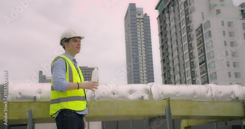 Young Caucasian engineer, wearing a hard hat and vest, hydrating with a water bottle on a building rooftop, overlooking urban skyline. Represents construction, safety, and modern development.