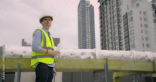 Young Caucasian engineer observing rooftop infrastructure, holding a water bottle amidst city buildings, symbolizing urban development, safety, and diligent maintenance work.