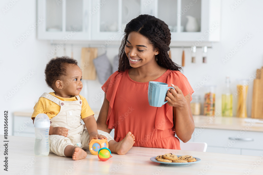 Fototapeta premium Portrait Of Happy Black Family Mom And Infant Son Spending Time In Kitchen At Home, Beautiful African American Mother Drinking Tea While Her Cute Toddler Boy Sitting On Counter Table, Free Space