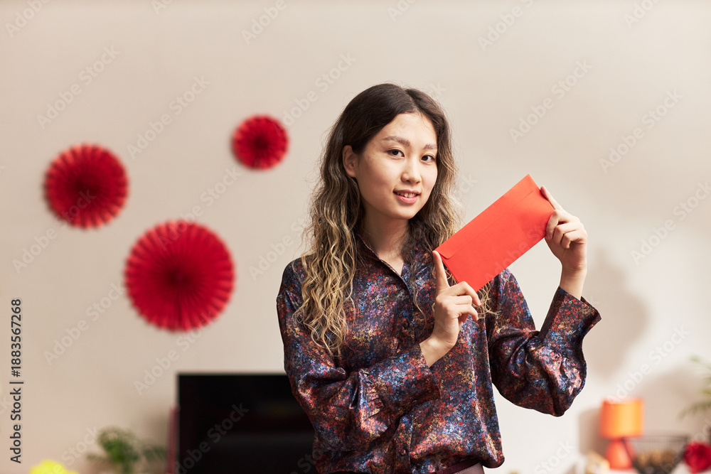 Fototapeta premium Portrait of young adult Asian woman smiling and holding red envelope with both hands, standing indoors with festive decorations in background during holiday celebration
