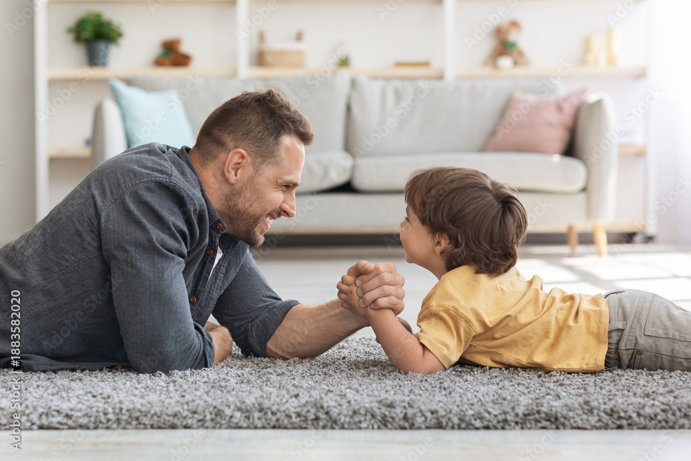 Fototapeta premium Bringing up strong kid. Happy father competing arm wrestling match with his little boy, lying together on floor at home and smiling to each other, profile portrait