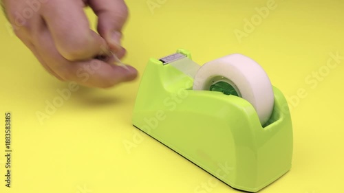 Hand cutting tape from a bright yellow dispenser on matching yellow background. Ideal for office, workspace, and organizational concepts. DIY work, school project, birthday present.